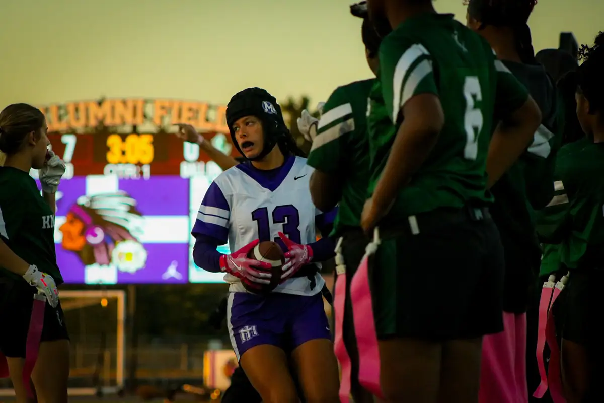 Flag football player in a purple number 13 jersey on the sideline at dusk, scoreboard glowing behind